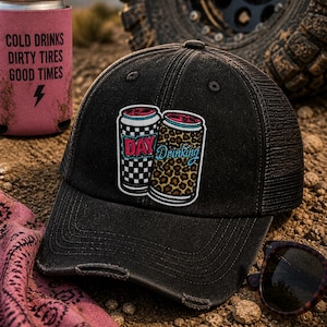 May include: Black trucker hat with a distressed brim, featuring a patch with two cartoon beverage cans. One can has a checkered pattern, the other a leopard print. The text "DAY Drinking" is on the patch. A pink can cooler with text is visible.