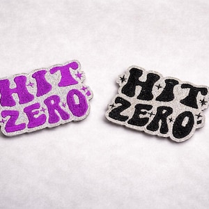 May include: Two embroidered patches with the text "HIT ZERO". One patch features purple text on a silver background, the other has black text on a silver background. Both patches have a decorative border.