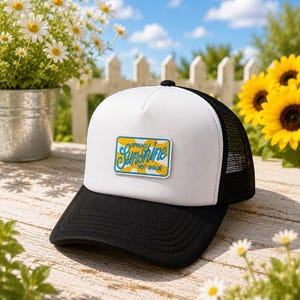 May include: A white and black trucker hat featuring a rectangular patch on the front panel. The patch displays a yellow sunburst graphic with the blue embroidered text "Spread Sunshine Not Shade" centered on the white foam front and mesh back.