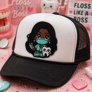 May include: Black and white trucker hat with a black brim and mesh back. The front panel is white with an embroidered patch of a cartoon dentist holding a dental tool and a tooth. The hat is a dental-themed accessory.