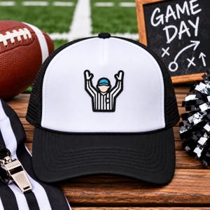May include: A white and black trucker hat featuring a centered embroidered patch of a referee with arms raised in a touchdown signal. The cap has a solid white front panel, a black brim, and black mesh back panels with a black cord trim detail.