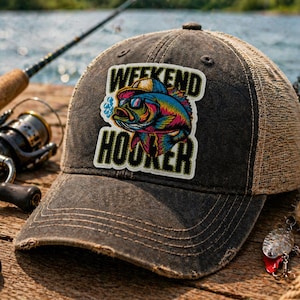May include: A distressed charcoal gray trucker hat with a mesh back. The hat features a patch with a colorful fish wearing sunglasses and the text "Weekend Hooker". Fishing gear is in the background.