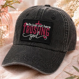 May include: A black, distressed baseball cap with a curved brim. The cap features a rectangular patch with the text "I'm just out here trusting God" in pink and white. The hat is made of a soft, worn material.