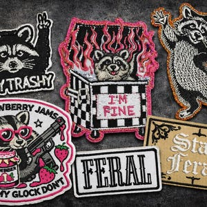 May include: A collection of embroidered patches featuring raccoons in various poses. One patch says "STAY TRASHY", another shows a raccoon in a dumpster with "I'M FINE". Other patches include "FERAL" and "Strawberry Jams but my Glock don't".
