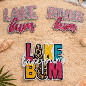 May include: Embroidered patches with the phrases "Lake Bum" and "River Bum" in pink and silver glitter, and a multi-colored "Lake Bum" patch with leopard print. The patches are displayed on sand with seashells and a starfish.