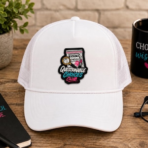 May include: A white trucker hat featuring a central patch with a graphic of an evidence bag, a magnifying glass, and a lipstick tube. The text on the patch reads "QUESTIONABLE CHOICES CLUB" in black and teal lettering with a pink accent.