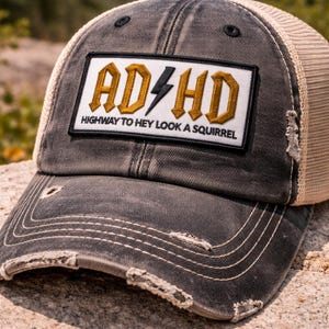 May include: A distressed gray trucker hat with a mesh back and a curved brim. The front patch features the text "AD/HD" in gold lettering with a lightning bolt, and the phrase "HIGHWAY TO HEY LOOK A SQUIRREL" below.