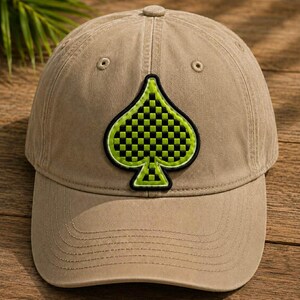May include: A tan baseball cap featuring a green and black checkered spade patch. The cap has a curved brim and a button on top. The spade design is outlined in black and green. The hat is made of a durable fabric.