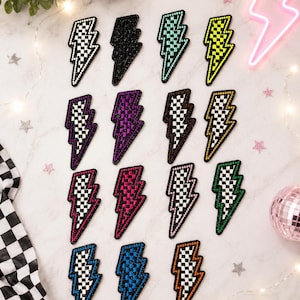 May include: A collection of sixteen lightning bolt patches arranged in a grid. Each patch features a checkered pattern in various colors including black, white, neon green, purple, pink, blue, orange, and green, all outlined with sparkling rhinestones.