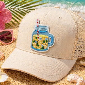 May include: Tan trucker hat with a mesh back and a patch of a lemonade jar with lemons and a red and white striped straw. The hat is on a sandy beach with pink sunglasses and a starfish.