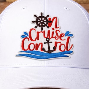 May include: White trucker hat with a mesh back and a curved brim. The front features an embroidered patch with the words "Cruise Control" in red script, along with a ship's wheel, anchor, and blue water design.