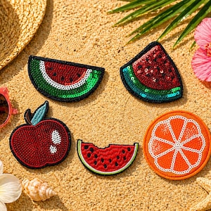May include: A collection of sequined fruit patches, including watermelon, apple, and orange slices, arranged on a sandy surface. A straw hat, sunglasses, and tropical flowers are also present, evoking a summer theme.