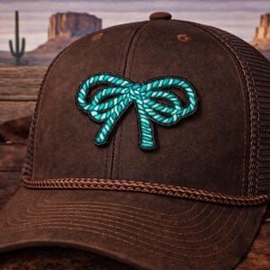 May include: A brown trucker hat with a turquoise rope bow patch. The hat has a mesh back and a decorative turquoise and gold pin on the side. The hat is set against a desert landscape.