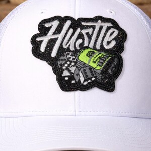 May include: White baseball cap with a mesh back and a curved brim. The cap features a patch with the word "Hustle" in white script, along with dice and a stack of money. The patch has a black glitter border.