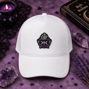 May include: A white trucker hat featuring a centered embroidered patch of a black cauldron with purple accents, white steam, and a triple moon symbol. The hat has a structured front panel and a mesh back, showcasing a clean, minimalist design.