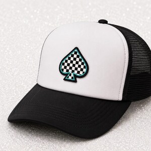 May include: A trucker hat featuring a solid black brim and a white front panel. Centered on the front is an embroidered spade symbol with a black and white checkered pattern, outlined in a vibrant teal border. The back panels are black mesh.