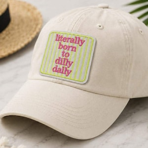 May include: A beige baseball cap with a curved brim. The cap features a square patch with the text "literally born to dilly dally" in pink letters on a striped background. A straw hat is partially visible in the background.