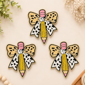May include: Three identical embroidered patches, each featuring a yellow pencil centered behind a decorative bow. The bow has a tan and black leopard print on the top loops and a white and black dalmatian-style spotted print on the bottom tails.