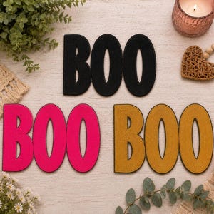 May include: Three sets of the word BOO in different colors. One set is black, one set is bright pink, and one set is mustard yellow. Each set features the letters B, O, and O connected together in a bold, sans-serif font with a textured surface.