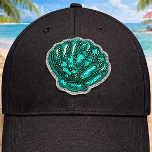 May include: Black baseball cap with a teal sequined seashell patch. The seashell is outlined with silver rhinestones. The cap has a curved brim and a fabric strap closure. The background shows a beach scene with palm trees and blue skies.