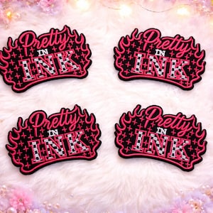 May include: Four embroidered patches with the text "Pretty in Ink" in a stylized font, outlined in pink and black. The patches have a flame design and a checkered pattern within the letters. The patches are on a white, fluffy surface.