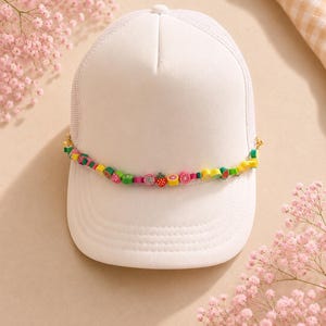 May include: A white trucker hat featuring a mesh back and a curved brim. A decorative string of colorful, small fruit-shaped beads, including strawberries, kiwis, and citrus slices, is attached across the front panel of the hat.