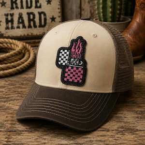 May include: A beige and brown trucker hat with a black and pink checkered lighter patch. The hat has a curved brim and a mesh back. The text "RIDE HARD" is visible in the background.
