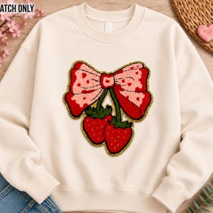 May include: A cream-colored crewneck sweatshirt featuring a large, textured patch on the chest. The patch depicts a pink bow with red heart patterns, attached to two bright red strawberries with green stems, all outlined in a gold-colored border.