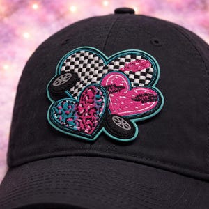 May include: Black baseball cap with a colorful heart-shaped patch. The patch features a checkered heart, a pink heart with black accents, and a teal leopard print heart, each with a small tire graphic.