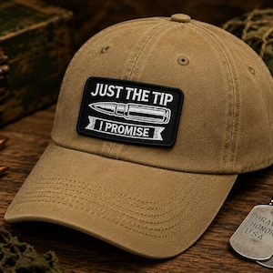 May include: A tan baseball cap with a black patch that reads "JUST THE TIP I PROMISE" in white text. The cap is on a wooden surface with two bullets and a military-style dog tag.
