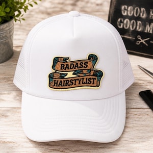 May include: A white trucker hat featuring a central embroidered patch with a tan ribbon design and dark teal accents. The text BADASS HAIRSTYLIST is stitched in black capital letters across the center of the patch. The hat has a solid front panel.