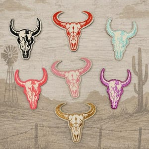 May include: A collection of embroidered bull skull patches in various colors, including black, red, light blue, pink, gold, and purple. The patches are arranged on a rustic background with a windmill, cactus, and barbed wire fence.