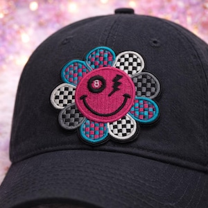 May include: A black baseball cap with a colorful flower patch. The flower has a pink center with a smiling face, an 8-ball eye, and a lightning bolt. The petals are checkered in black, white, and gray, and teal and pink.