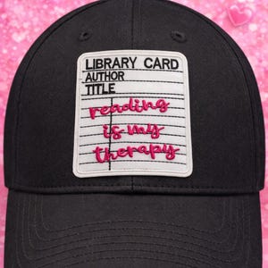 May include: Black baseball cap with a white patch featuring the words "LIBRARY CARD AUTHOR TITLE" above the phrase "reading is my therapy" in pink script. The cap has a curved brim and a button on top.