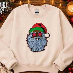 May include: A beige crewneck sweatshirt featuring a central embroidered patch of a Santa Claus figure wearing red-tinted circular sunglasses and a red and green hat. The Santa has a textured light blue beard and the text PATCH ONLY is visible.