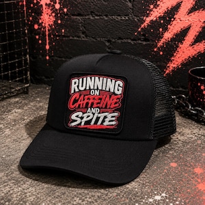 May include: A black trucker hat featuring a rectangular patch on the front. The patch displays the white and red embroidered text RUNNING ON CAFFEINE AND SPITE. The hat has a solid front panel and a mesh back panel with an adjustable snap closure.