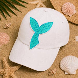 May include: White baseball cap featuring a turquoise mermaid tail design. The cap has a curved brim and a classic six-panel construction. The mermaid tail is a vibrant turquoise color with a scale pattern.