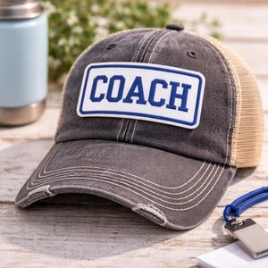 May include: A distressed gray baseball cap with a tan mesh back. The front features a white rectangle with the word "COACH" in blue block letters. The cap has a pre-curved bill and a distressed look.