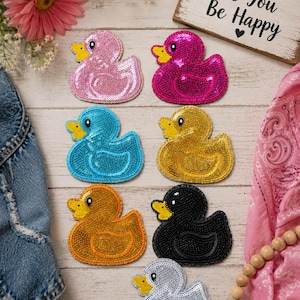 May include: A collection of sequined duck patches in various colors, including pink, magenta, blue, gold, orange, black, and silver. Each duck has a yellow beak and is arranged on a light wooden surface. A sign reads "Be Kind Be You Be Happy."