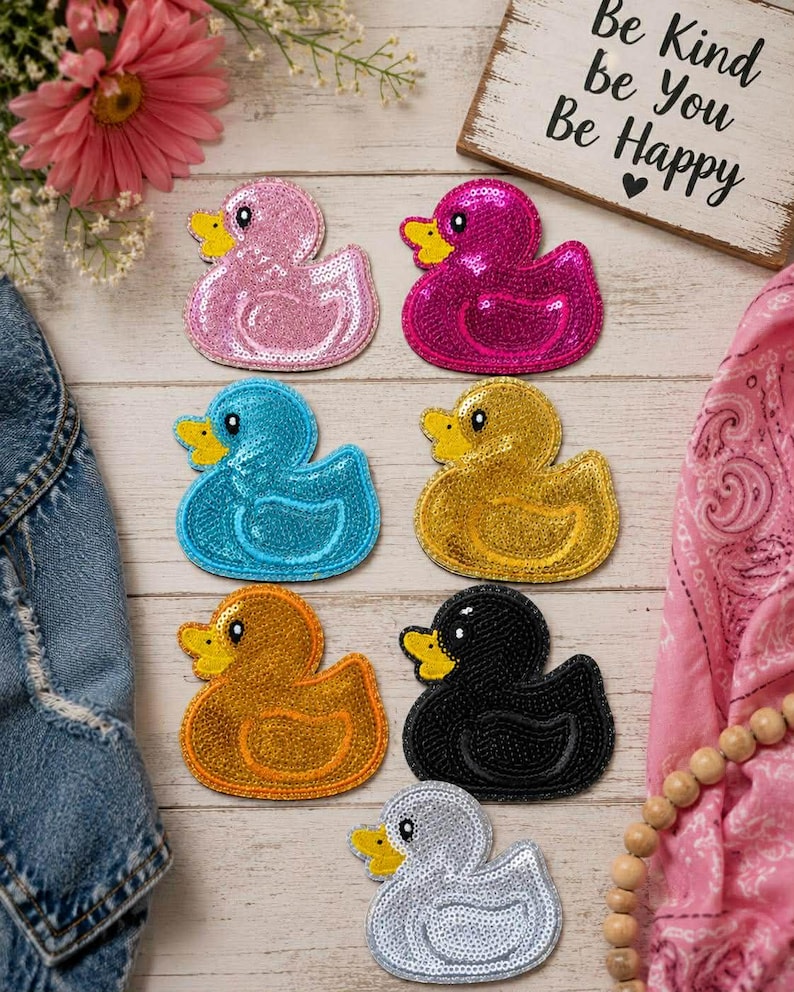 May include: A collection of sequined duck patches in various colors, including pink, magenta, blue, gold, orange, black, and silver. Each duck has a yellow beak and is arranged on a light wooden surface. A sign reads "Be Kind Be You Be Happy."