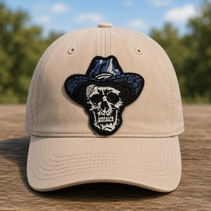 May include: A tan baseball cap with a black and blue patch featuring a skull wearing a cowboy hat. The hat has a curved brim and a button on top. The patch is centered on the front of the cap.