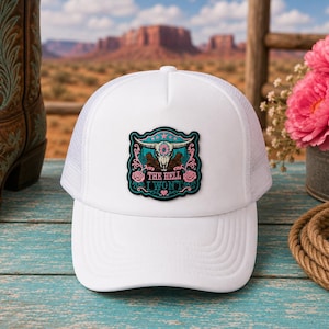 May include: A white trucker hat featuring a central patch with a steer skull, floral accents, and the text THE HELL I WON'T. The cap has a solid front panel and a mesh back, showcasing a detailed graphic design with pink, teal, and black colors.