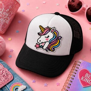 May include: A black and white trucker hat featuring an embroidered unicorn patch with a pink, purple, and yellow mane, a gold horn, and a pink heart cheek. The hat has a solid white front panel, a black brim, and black mesh back panels.