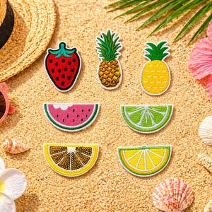 May include: A collection of embroidered fruit patches, including a strawberry, pineapple, watermelon, lime, and lemon, arranged on a sandy surface. A straw hat, sunglasses, and a pink hibiscus flower are also present, evoking a summer theme.