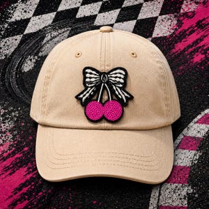 May include: Tan baseball cap with a cherry and bow patch. The patch features a black and white bow above two pink cherries. The cap has a curved brim and a button on top. The background has a checkered pattern and pink accents.