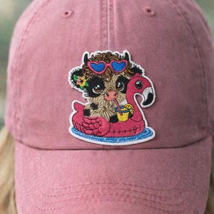 May include: A dusty rose baseball cap with a colorful embroidered patch. The patch features a cartoon cow wearing heart-shaped sunglasses and a flower crown, floating on a pink flamingo pool float with a drink.