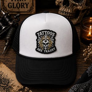 May include: A black and white trucker hat featuring a central embroidered patch. The patch displays a skull, gold roses, and spiderwebs with the text "TATTOOS ARE TRASHY" in a bold, arched font. The hat has a white front panel and black mesh back.
