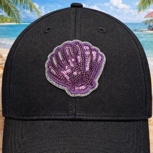 May include: Black baseball cap with a seashell motif. The shell is decorated with purple sequins and a silver glitter border. The cap has a traditional six-panel construction and a curved peak.