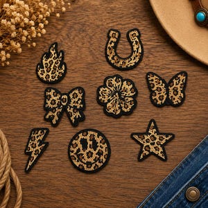 May include: A collection of eight embroidered patches featuring a tan and black leopard print pattern. The shapes include a flame, a horseshoe, a bow, a flower, a butterfly, a lightning bolt, a smiley face, and a five-pointed star with black borders.