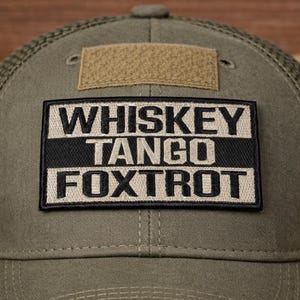 May include: Olive green trucker hat with a mesh back and a patch that reads "WHISKEY TANGO FOXTROT" in bold white letters on a black and tan background. The hat has a curved brim and a small tan square patch.